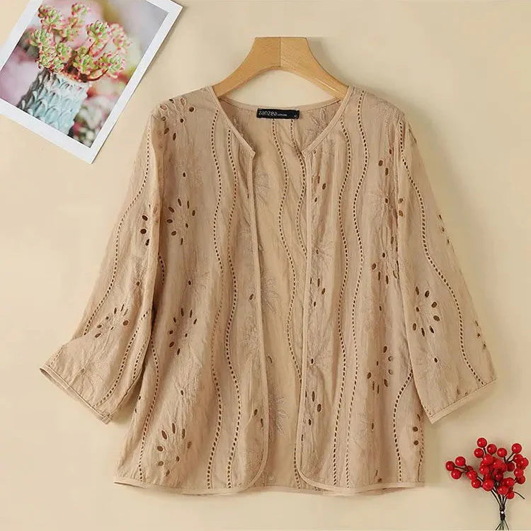 Beige embroidered women’s blouse with 3/4 sleeves, scalloped open front, and floral cut-out patterns. Displayed on a wooden hanger against a neutral background.